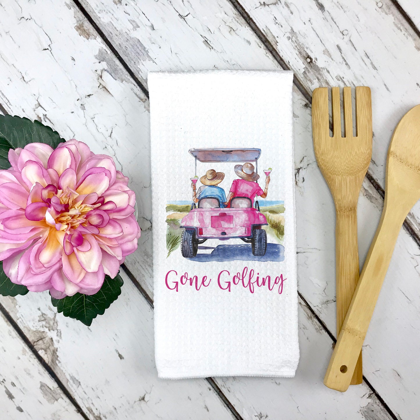 White waffle weave towel with a golf cart design and pink flower in left side of image, Gone Golfing phrase, two people in a golf cart