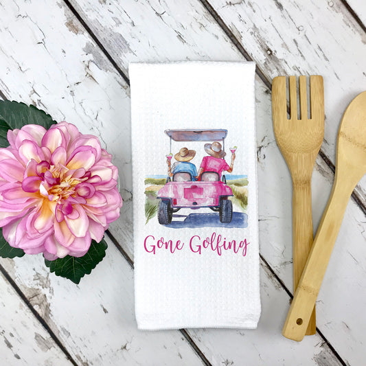 White waffle weave towel with a golf cart design and pink flower in left side of image, Gone Golfing phrase, two people in a golf cart